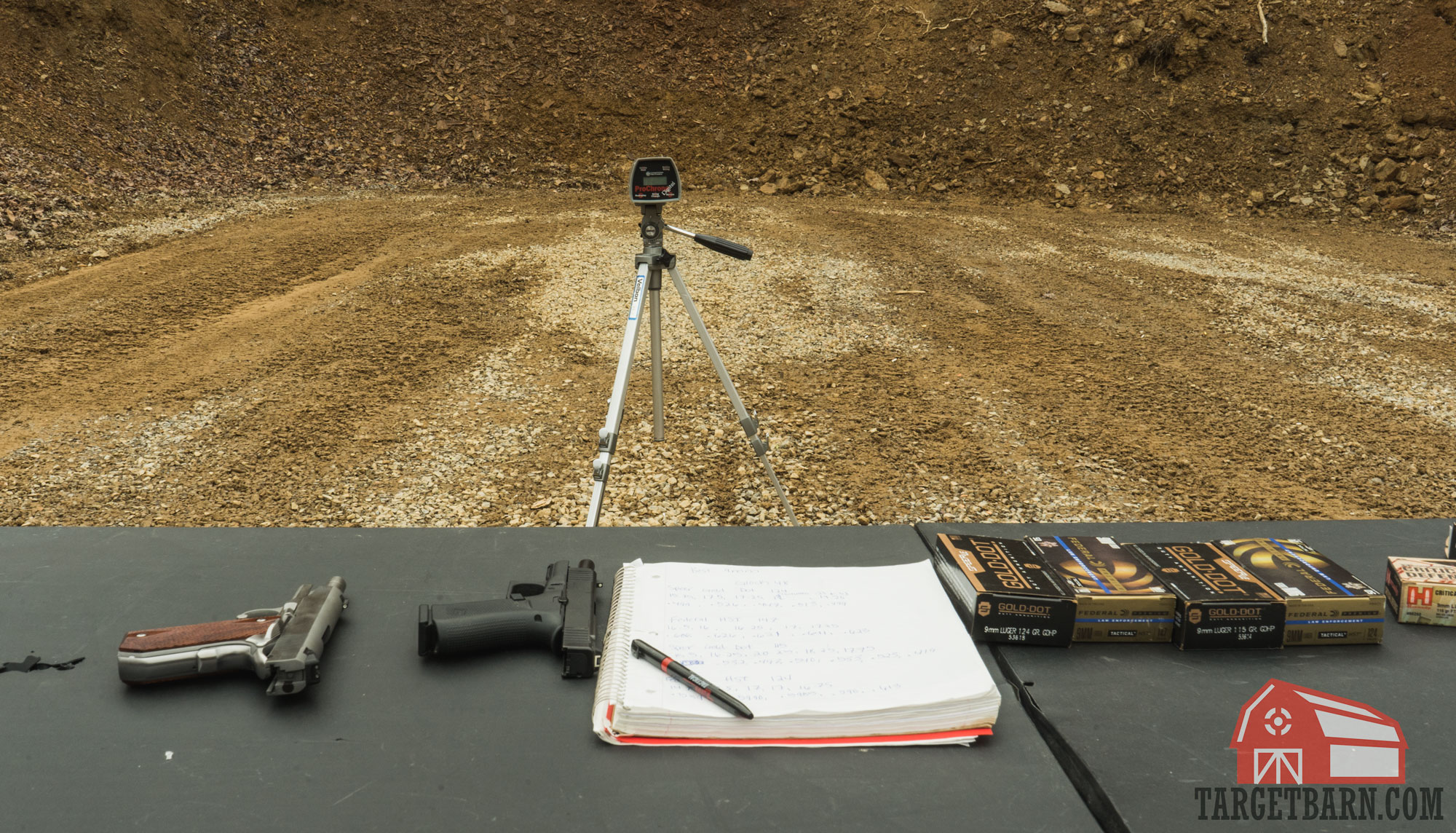 a table set up at the range with pistols, a notebook, and ammo boxes, and a chronograph set up down range to measure velocities